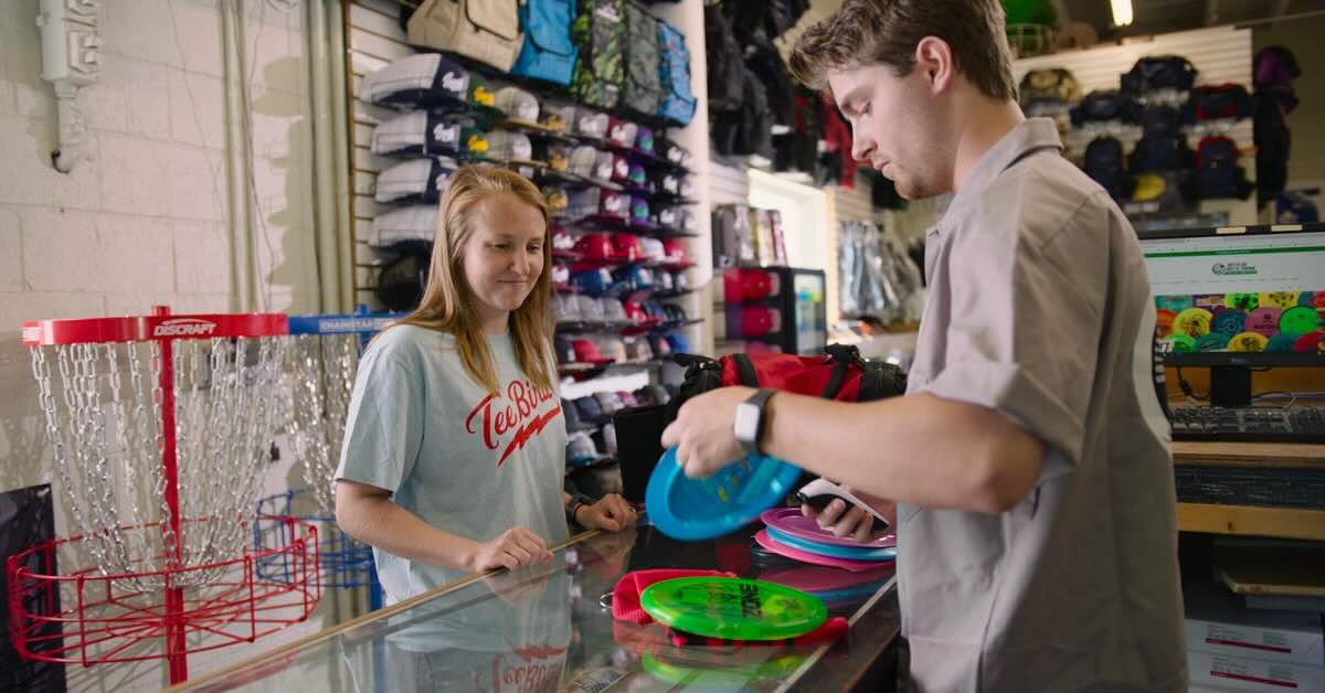 A man at a counter shows disc golf discs to a woman and appears to be explaining them to her