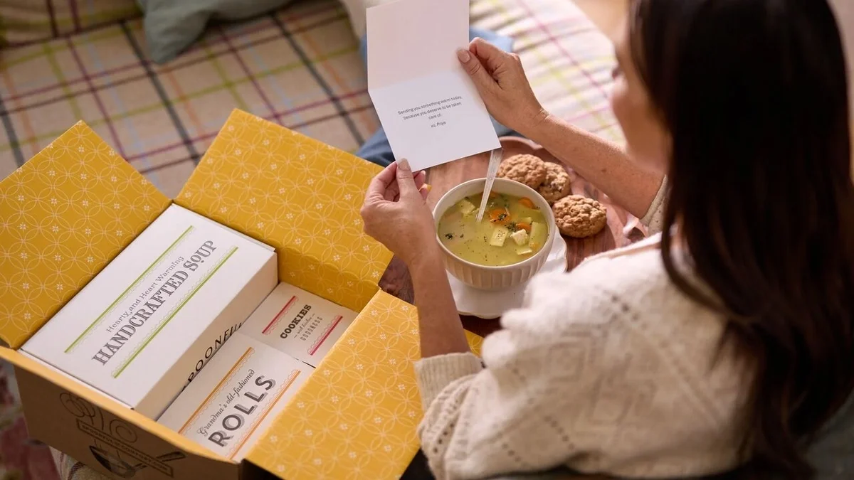 Overhead view of a woman reading a greeting card while enjoying a bowl of chicken soup and cookies, with an open Spoonful of Comfort gift box containing handcrafted soup and rolls beside her.