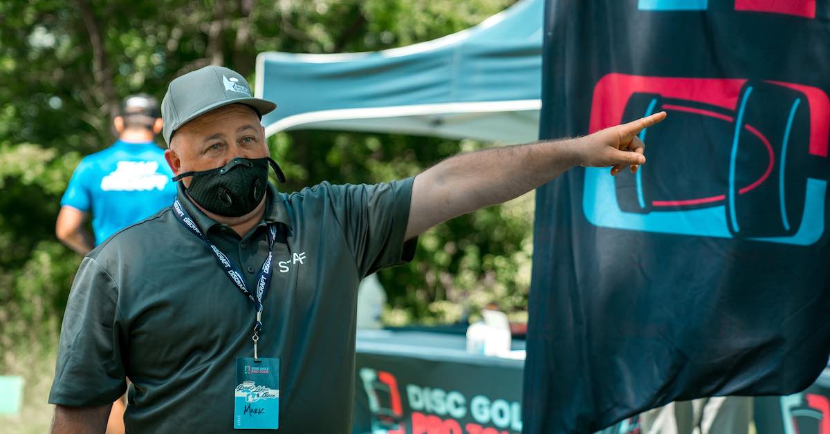 A man in a dry-fit polo and baseball cap points at seomthing while wearing a mask