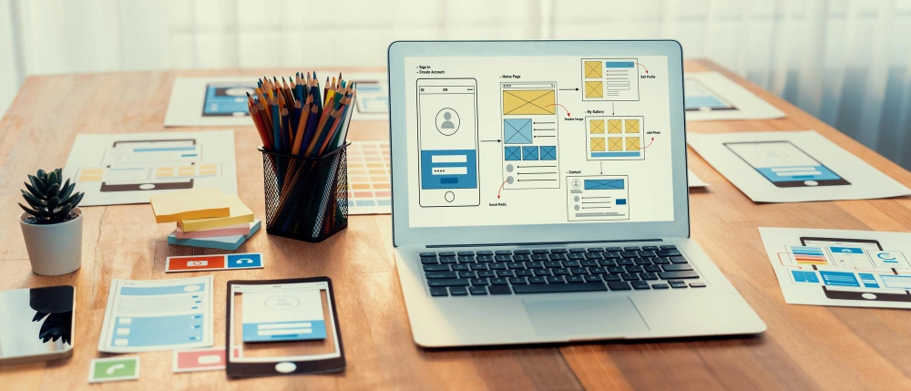 A photograph of a laptop on a desk displaying a wireframe design, surrounded by mobile devices and interface sketches, illustrating a user experience design process.