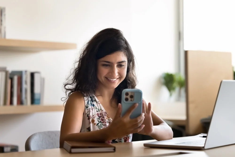A woman sat at her desk is checking her credit score on her mobile phone and smiling