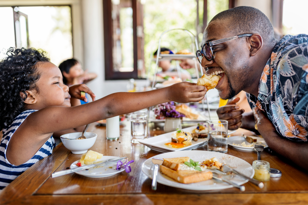 A daughter is feeding her father sandwiches in a cafe