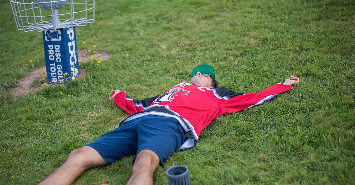 A man lays on the ground as if exhausted next to a disc golf basket
