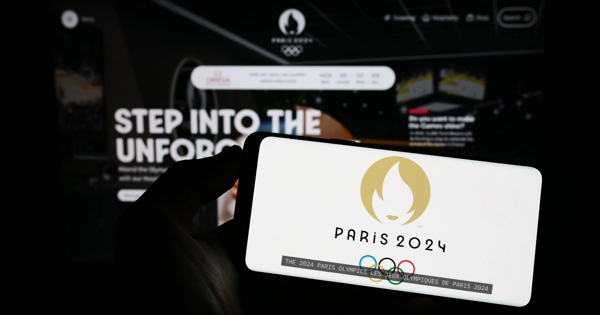 A cellphone displays the Paris 2024 Olympic logo with captions stating the same. In the background a website for the olympics is shown.