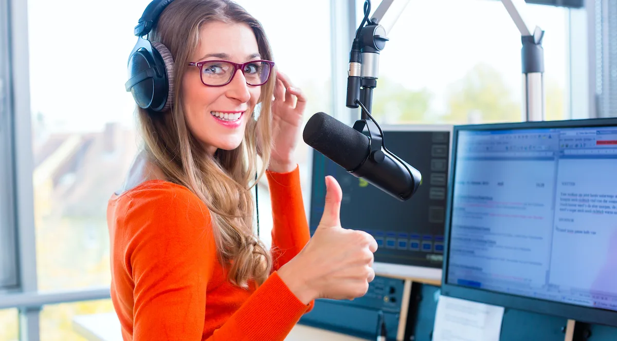 A woman sitting in front of a microphone with text on her screen, giving a thumbs up.