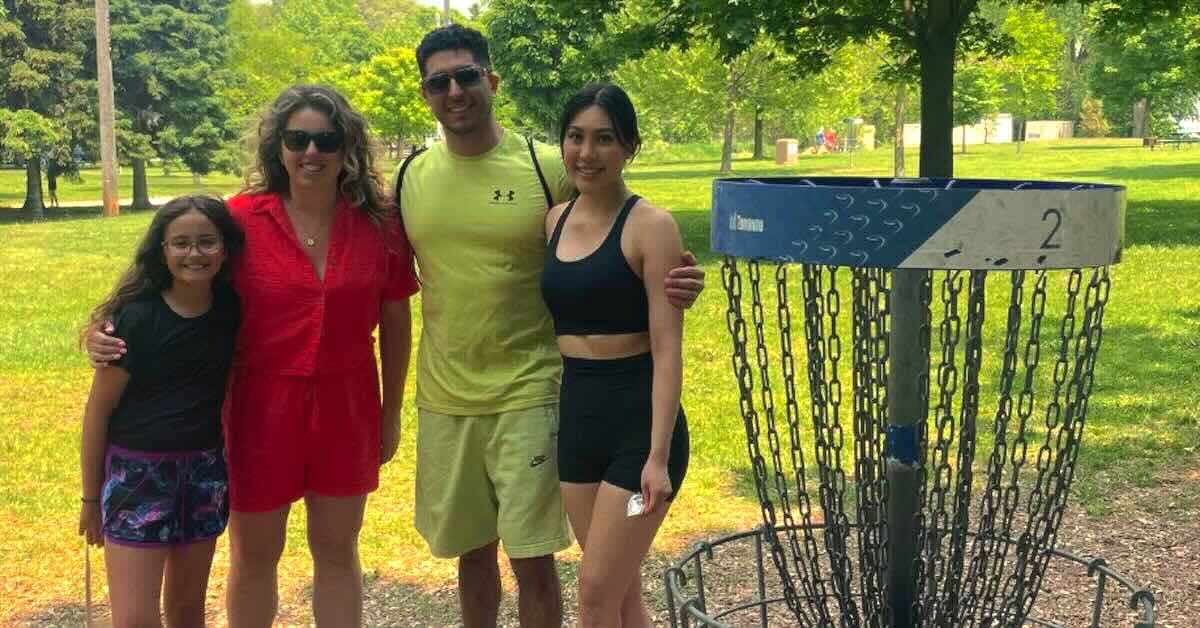 A family with one young girl, one young woman, an older woman, and a man pose for a photo near a disc golf basket