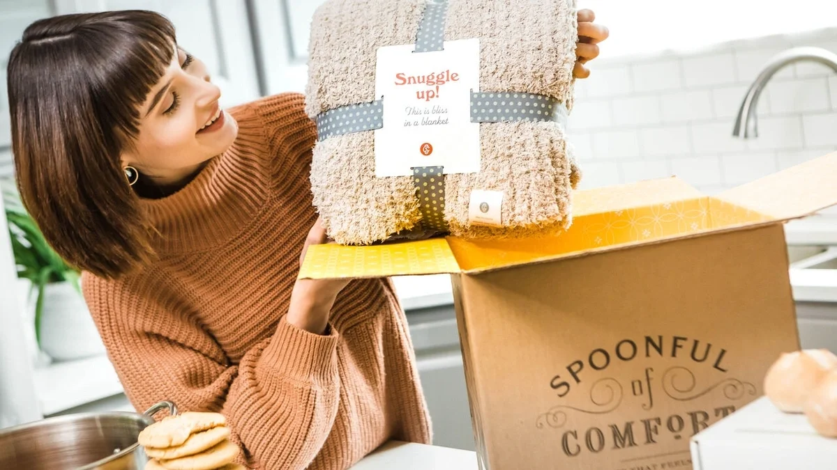 Woman smiling as she lifts a cozy throw blanket labeled “Snuggle Up” from an open Spoonful of Comfort gift box, with cookies and a bowl of soup nearby.