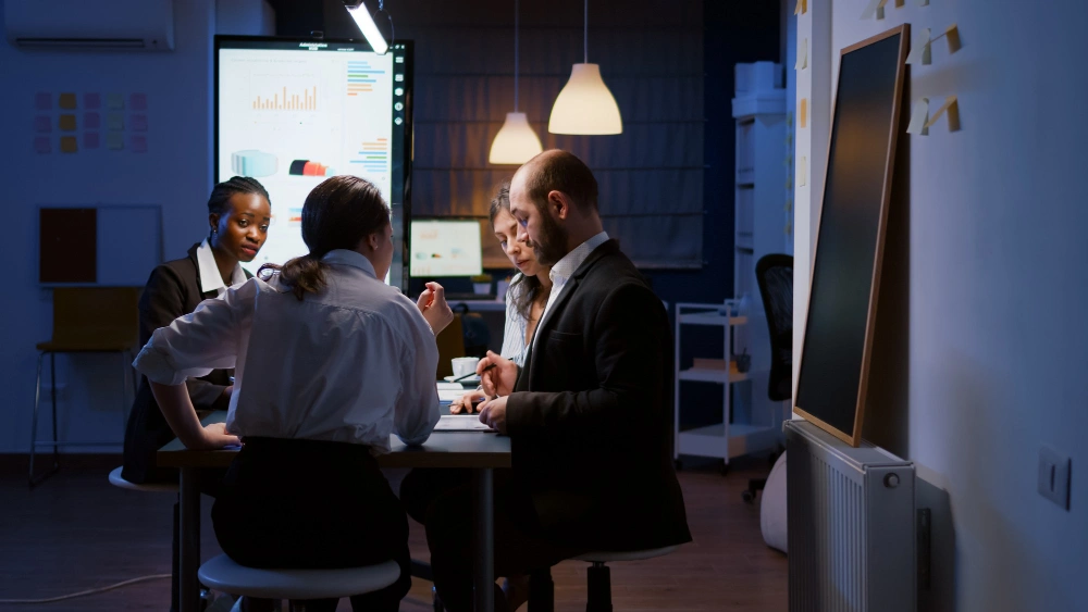 A diverse group of business professionals having a meeting in a dimly lit office with data charts displayed on a screen in the background.