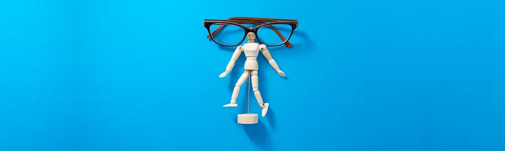 Image shows a miniature posable wooden figure-model, posed arms-open on a plain blue background. Over the head rests a folded women's reading glasses with brown frames.