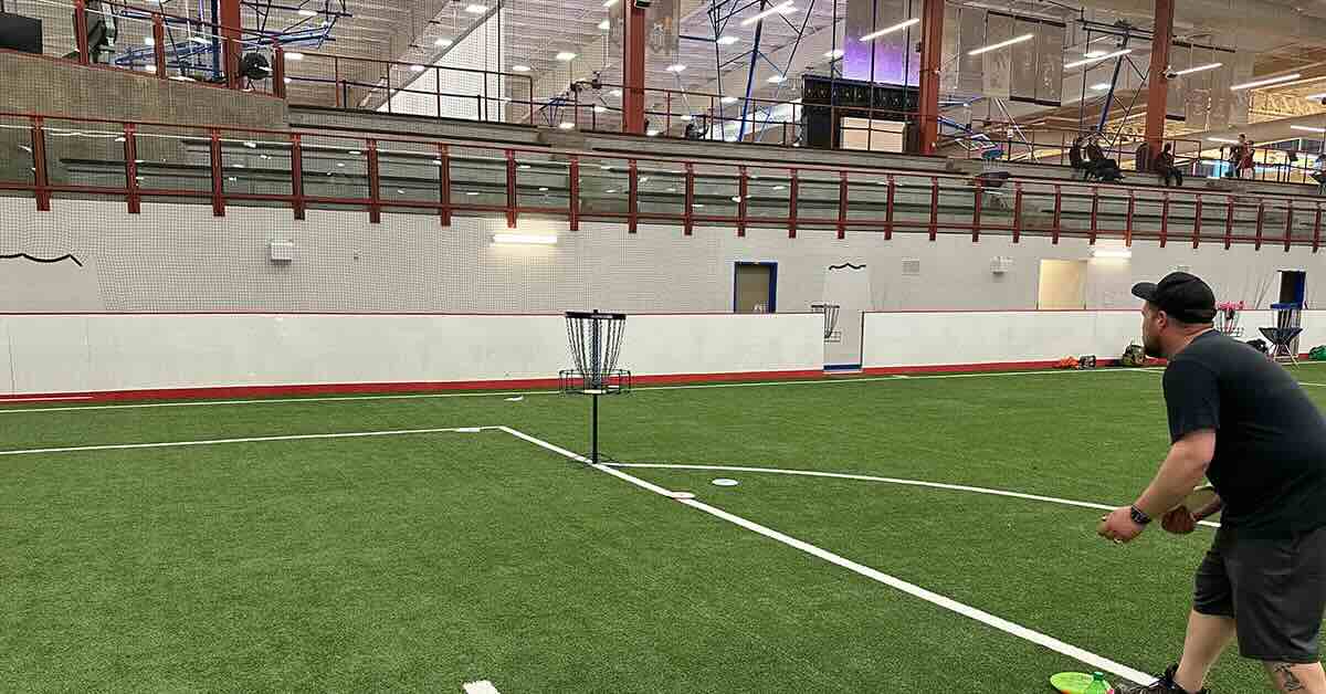 A man putts at a disc golf basket on an indoor soccer field