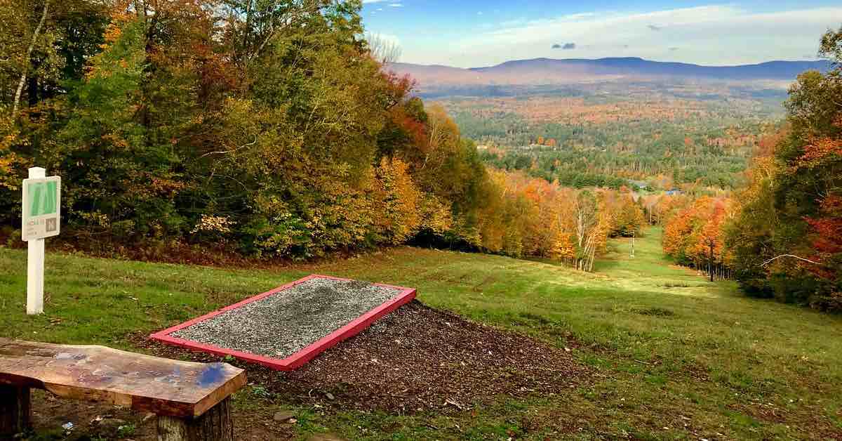 A disc golf tee pad at the top of a smooth, grassy slope with a view of trees in fall colors and mountains in the distance