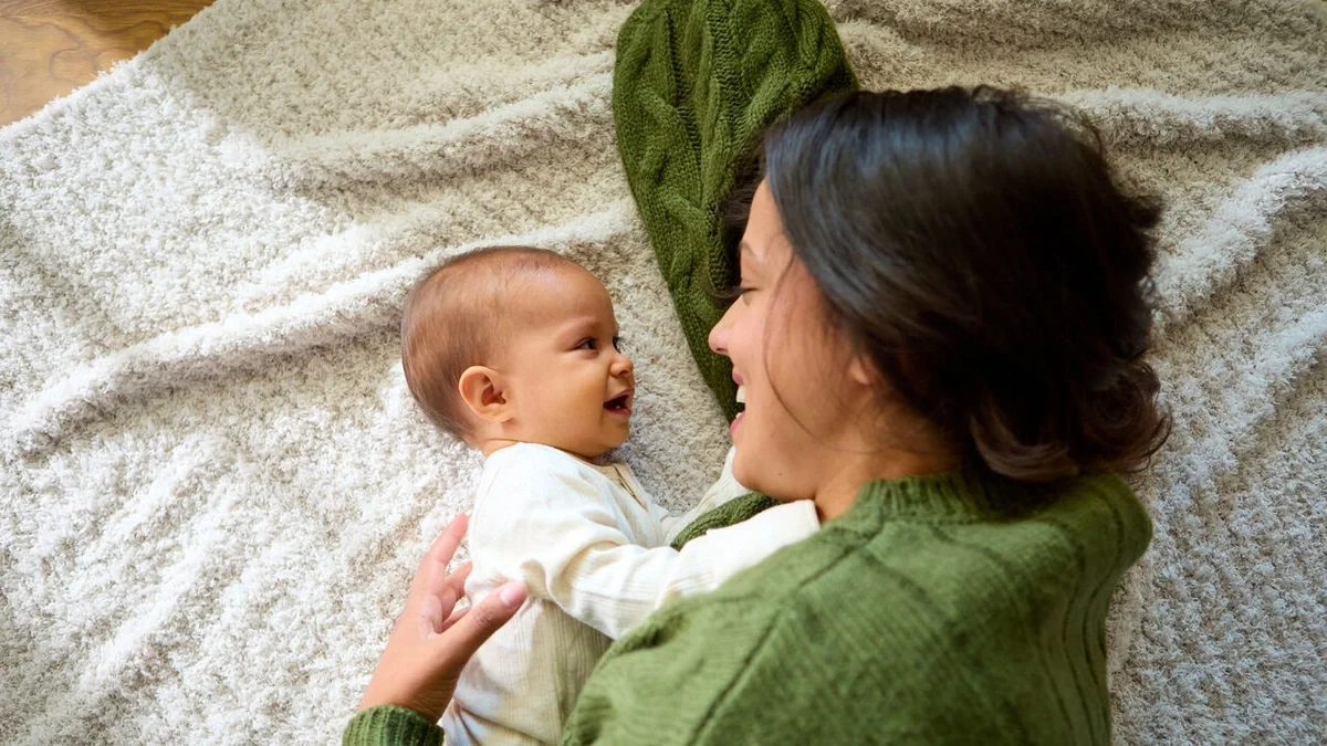 Parent lying on a soft blanket, smiling and cuddling with a happy baby as they look at each other.