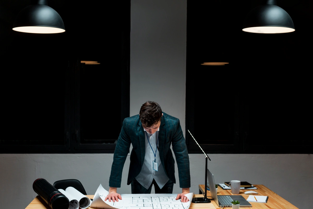 A young man in a suit standing over a desk covered with blueprints and documents, working late at night under two hanging lamps.