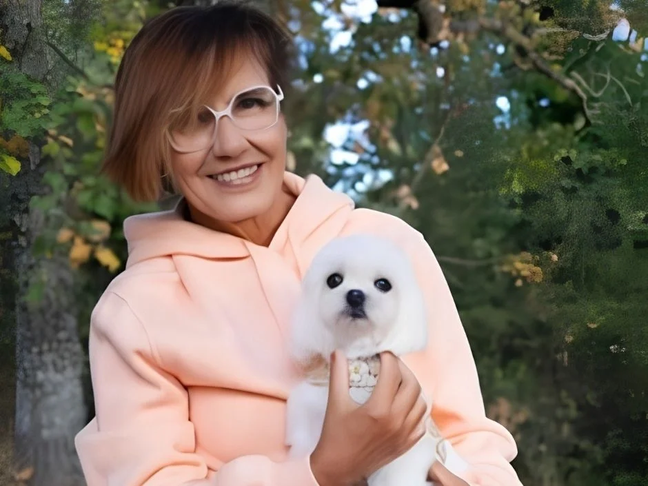 Middle-aged woman holding a tiny white Maltese puppy outdoors with trees in the background