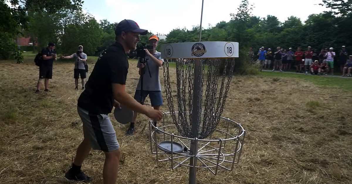 A crowd watches as a disc golfer in a black shirt gets a disc out of a basket while smiling