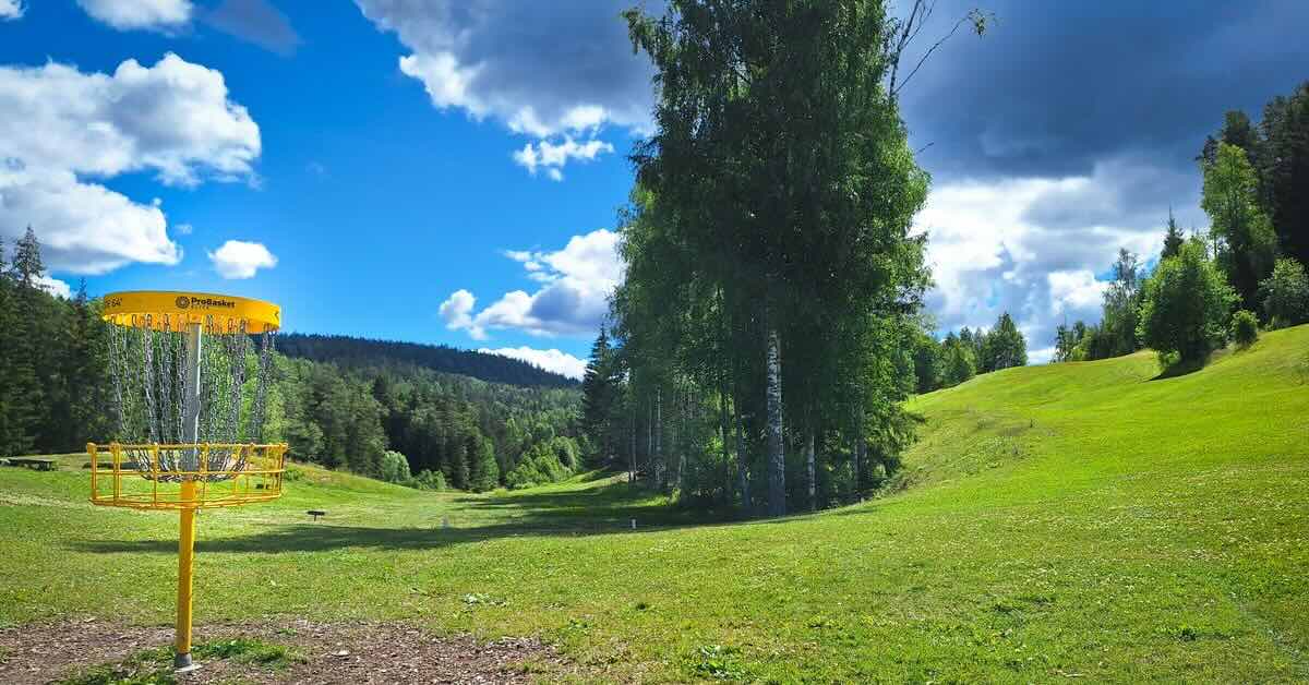 A yellow disc golf basket in an area of mown grass lined with trees and a hilly landscape