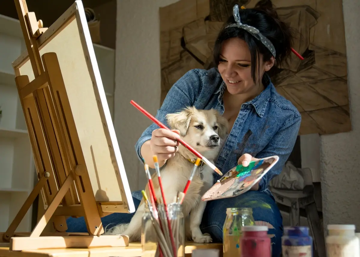 A dog and woman paint a picture together using a paint artist palette and easel