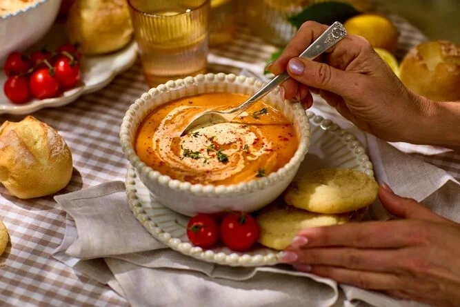 Styled image of tomato soup surrounded by tomatoes, lemon cookies and rolls