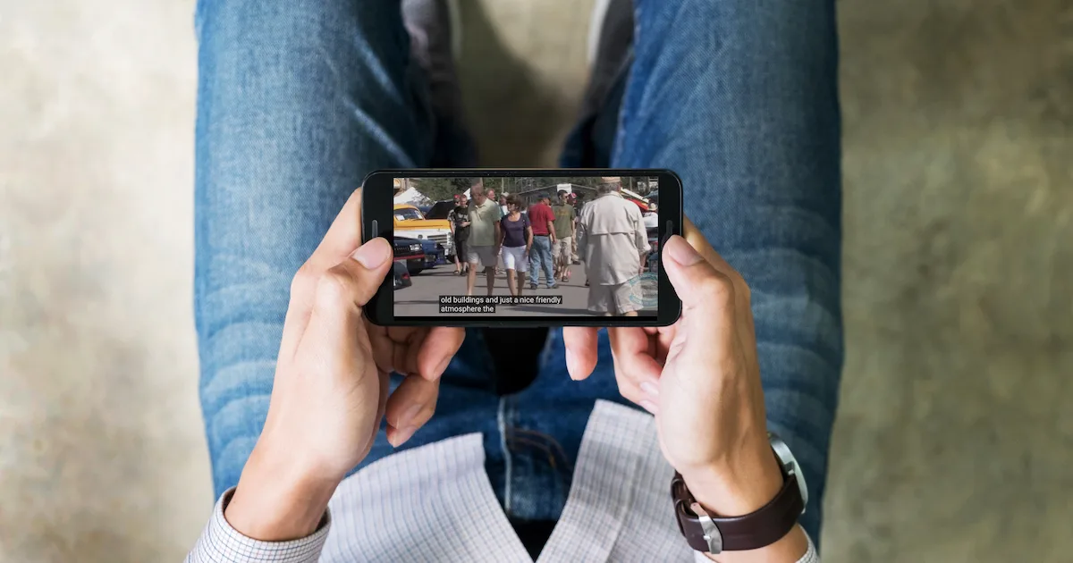 A person looking down from their point of view at a mobile phone in their hands which is displaying a video featuring closed captions.