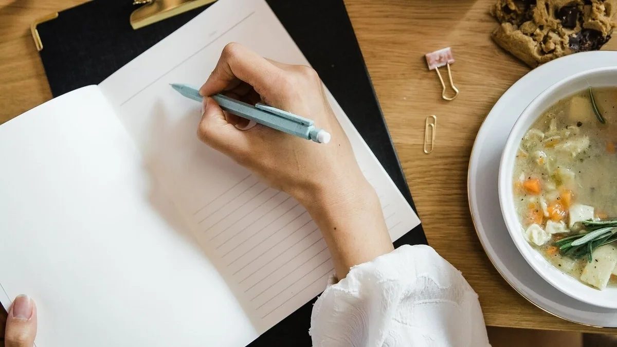 Overhead view of a person writing a handwritten note at a desk, with a bowl of chicken soup, a cookie, and office supplies nearby.