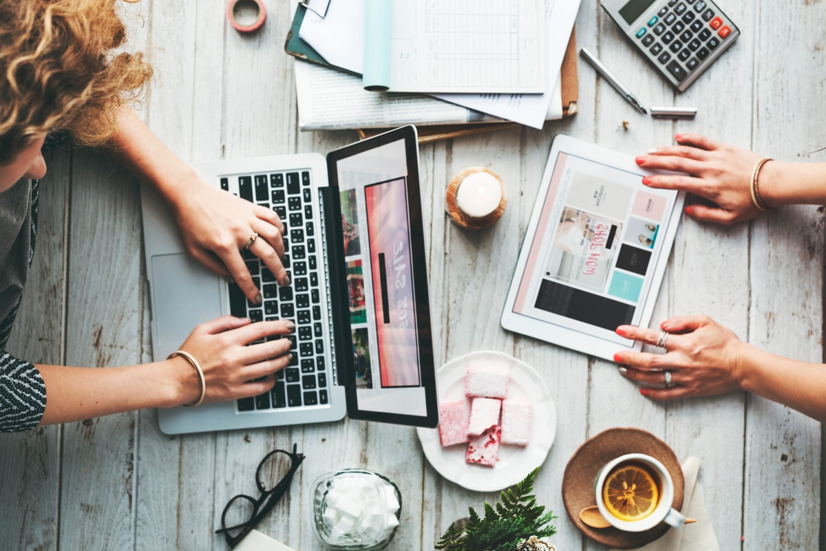 Two women on a computer and laptop doing work with coffee, papers, glasses, and more