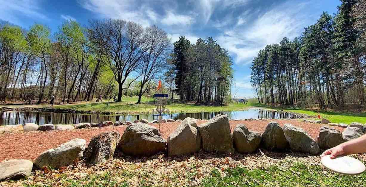 Shot of a disc golf green covered in red mulch and surrounded by large rocks with a small pond and trees also in view