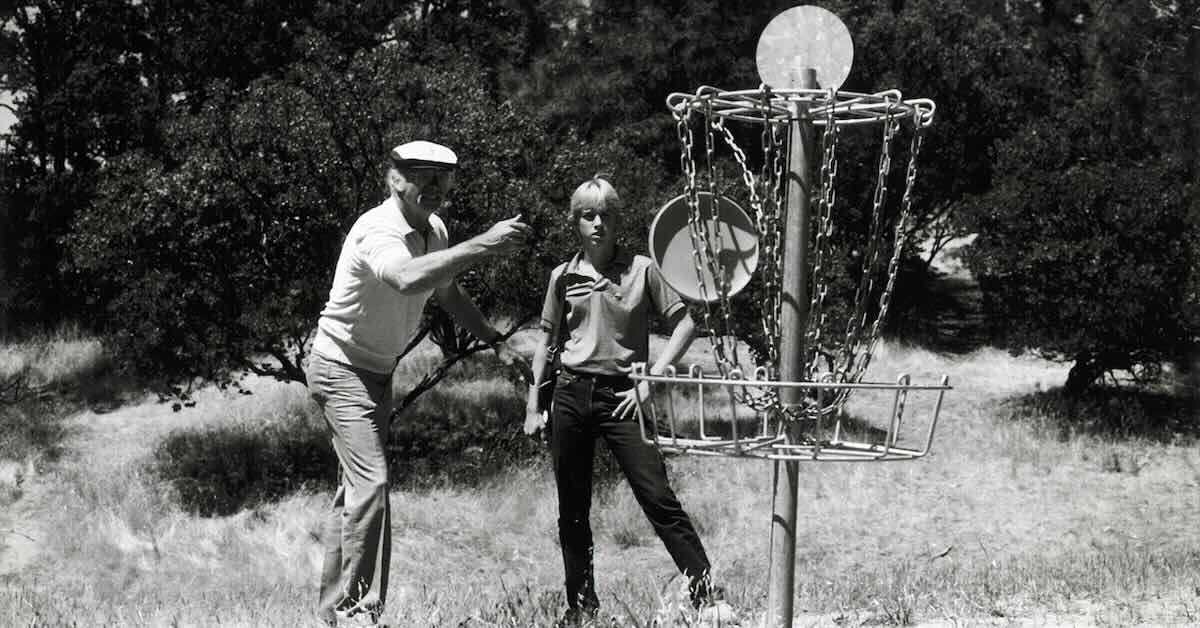 Black and white photo of an older man throwing a Frisbee into an original disc golf basket as a young man observes