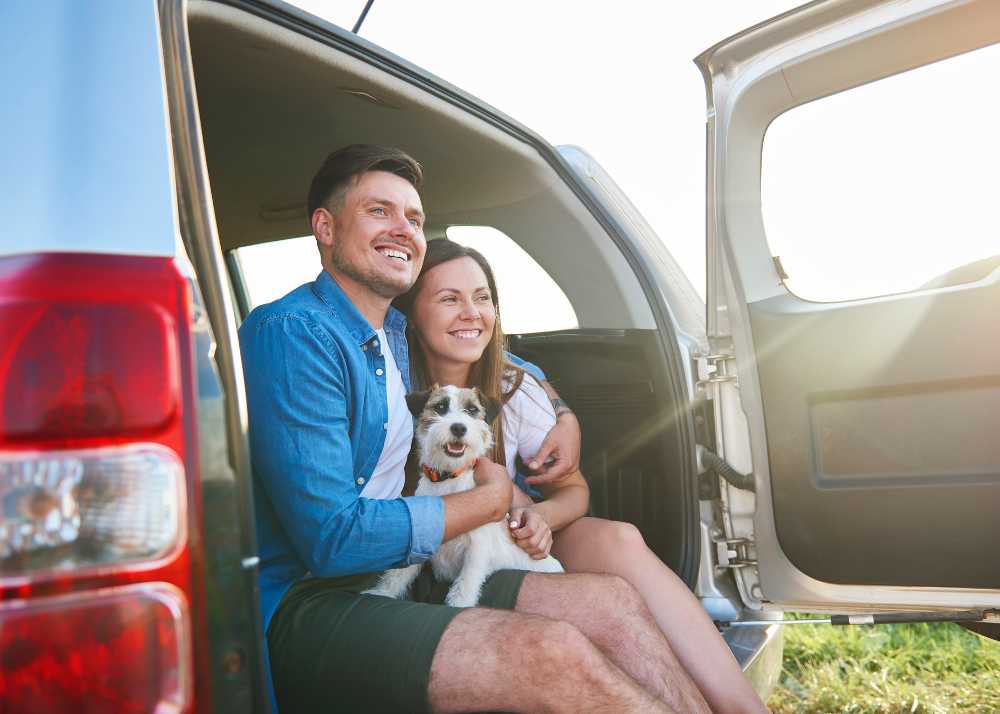 a man holding a dog and a woman sit in the back of a van with the gate open