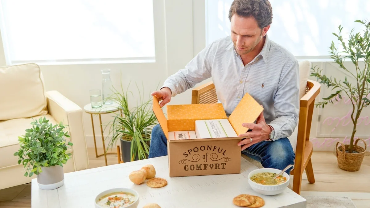 Man sitting in a bright living room opening a Spoonful of Comfort gift box, with bowls of soup, rolls, cookies, and potted plants on the coffee table in front of him.