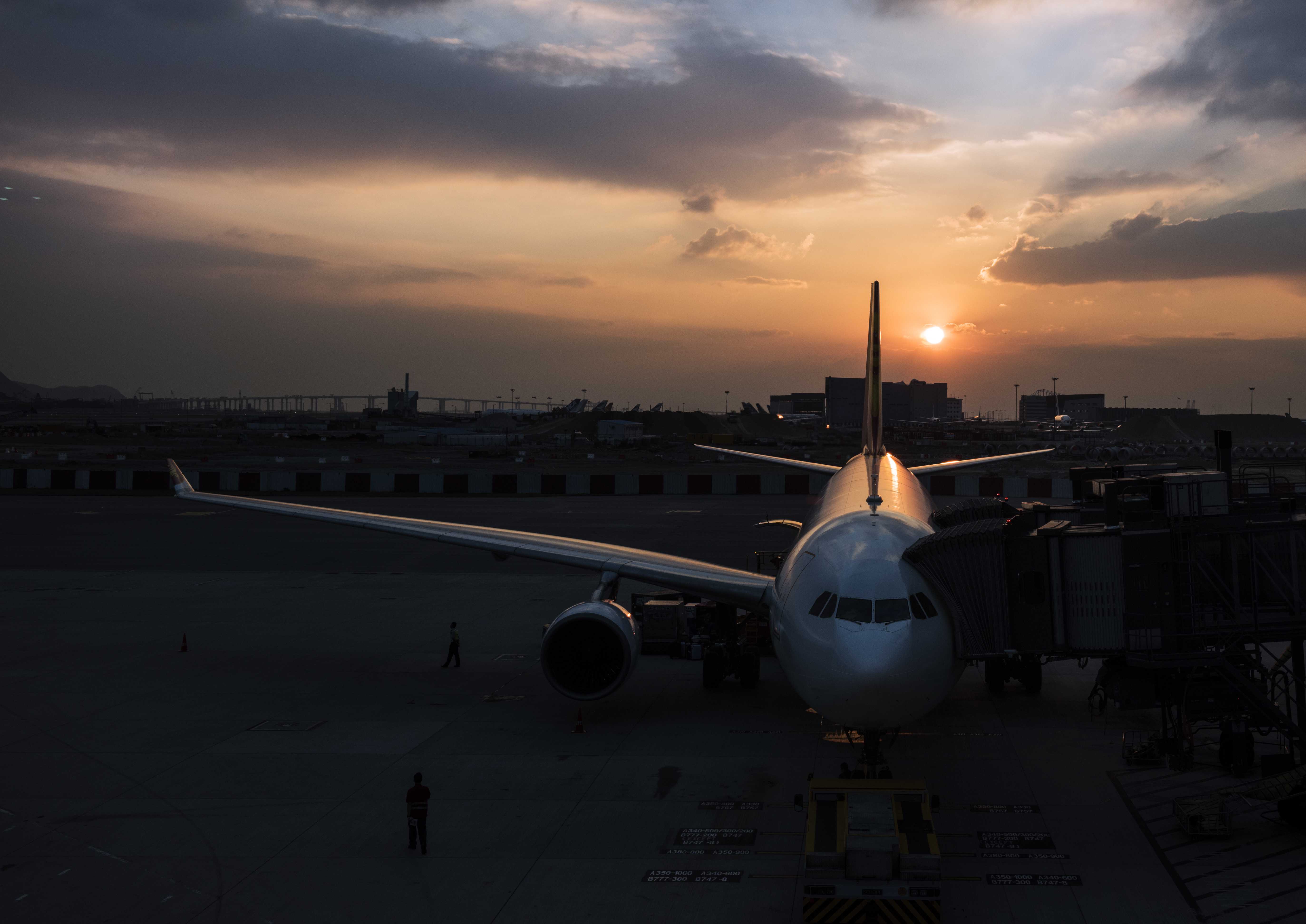 An airplane parked at an airport gate during sunset, with a dramatic sky and a runway in the background.