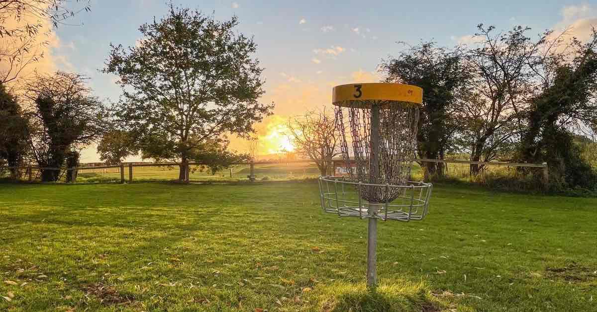 A disc golf basket in a field with the run rising behind it