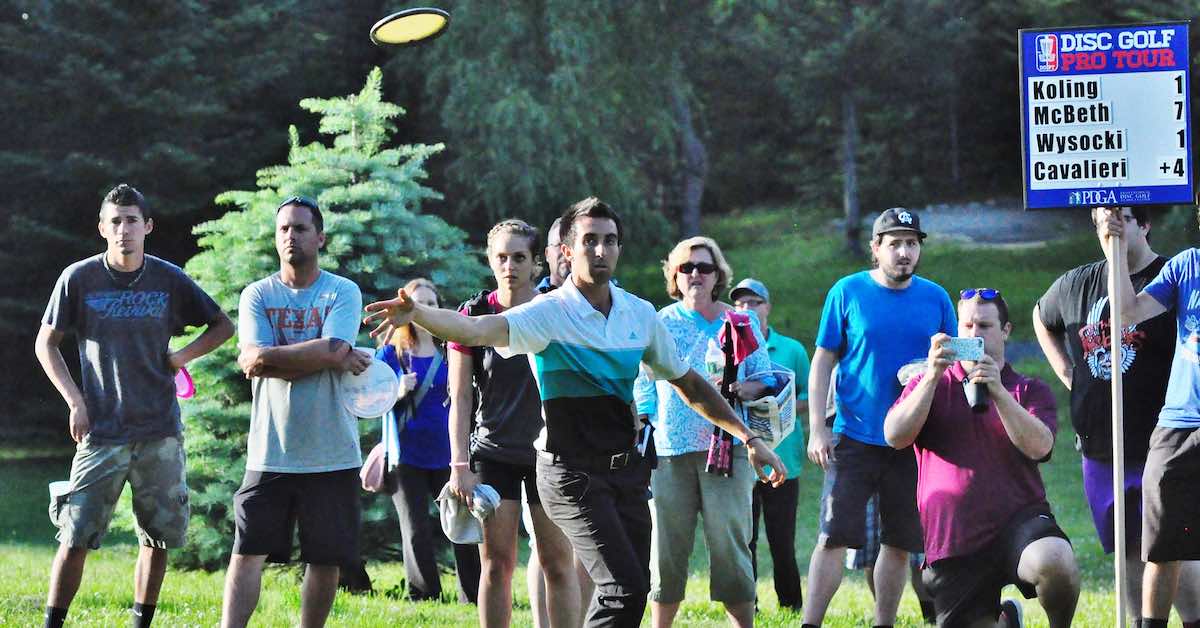 A young man throws a disc as a crowd looks on and a scoreboard saying "Disc Golf Pro Tour" at the top is in the background