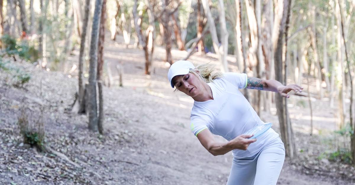 A woman dressed in white reaching back to throw a disc golf frive down a wooded fairway
