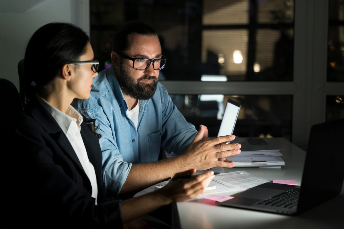 Two business professionals, a man and a woman, working late at an office desk with a laptop, documents, and a tablet, engaged in a serious discussion.