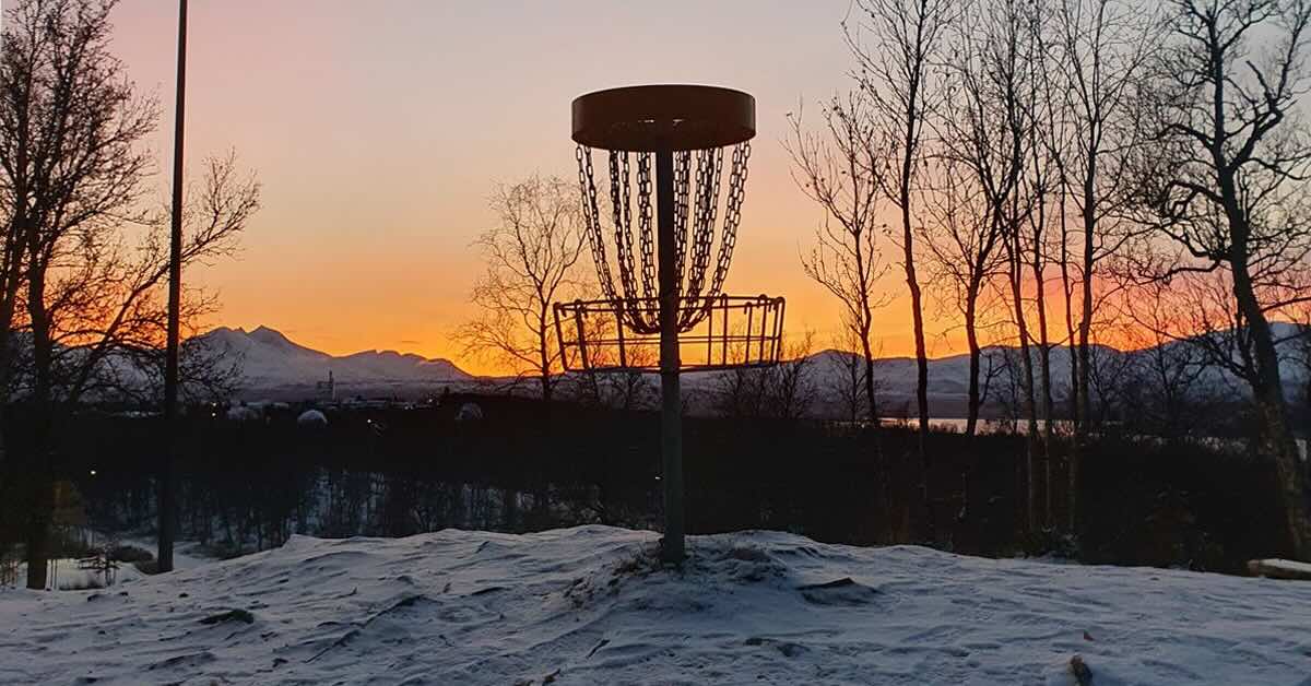 A disc golf basket on snowy ground on with mountains and a sunset in the background