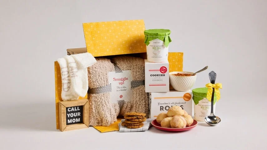 Comfort gift set displayed with a cozy throw blanket, white socks, two containers of soup with green lids, a box labeled “Rolls,” a box of cookies, rolls, cookies, a bowl of soup, a metal ladle with a yellow ribbon, and a wooden sign that reads “Call Your Mom.”