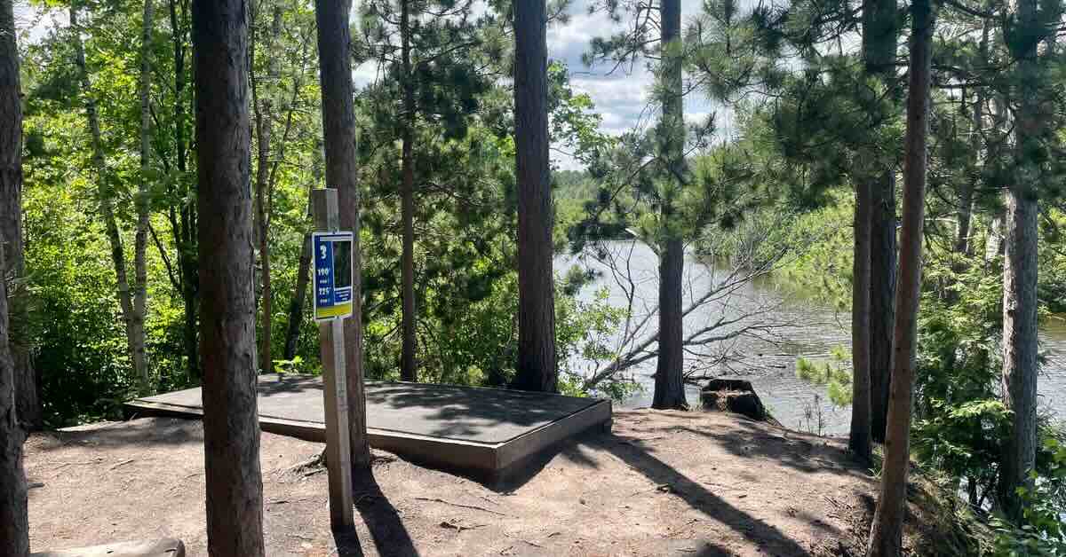 A rubber mat disc golf tee pad surrounded by pines with a river below in the background