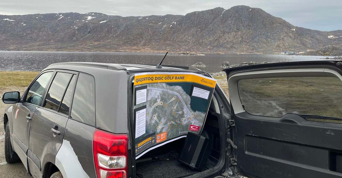 An SUV with it's back open and a disc golf course map hanging down. Water and bare, stone mountains in the distance