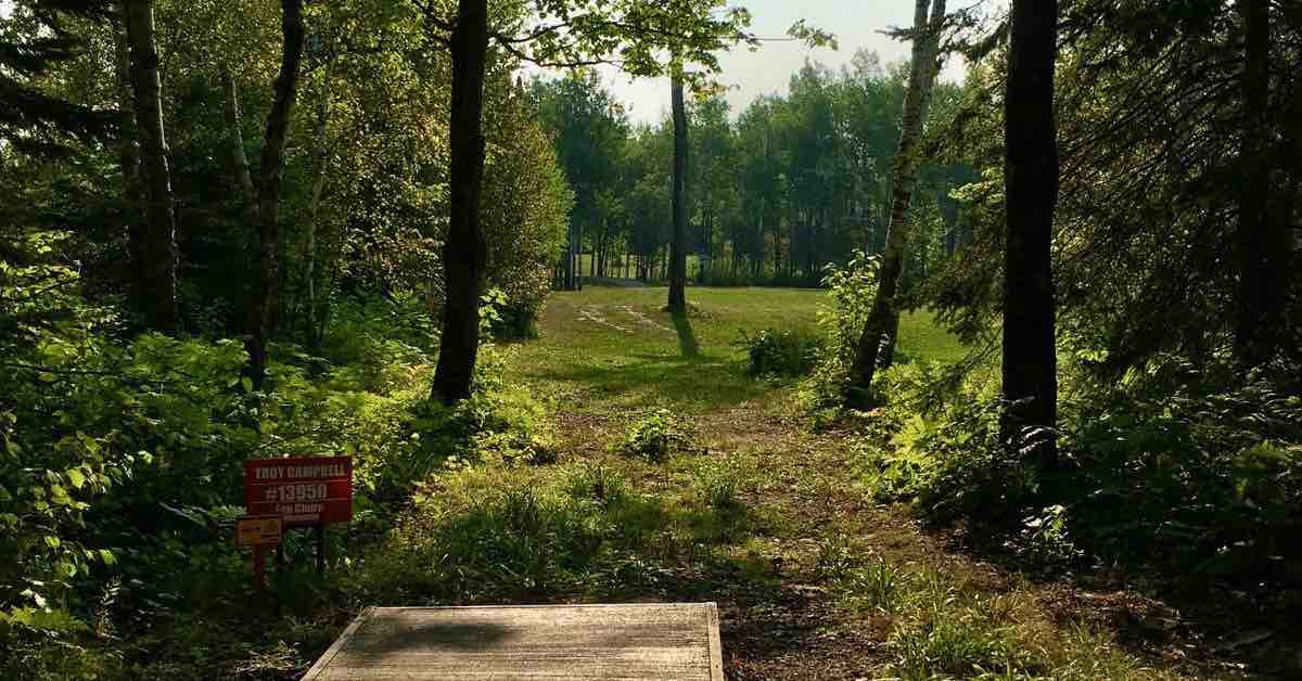 A concrete disc golf tee pad in a wooded area pointing to an open, mown field