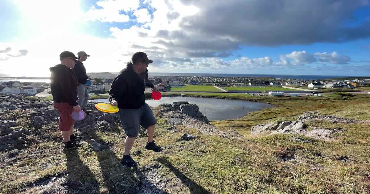 A man lining up a forehand disc golf throw watched by two other men on an elevated, rocky, treeless area with a view of town and sea beyond