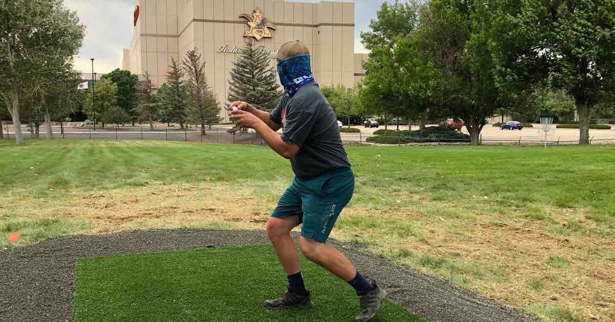 Disc golfer running up on tee pad in with large building and parking lot in background