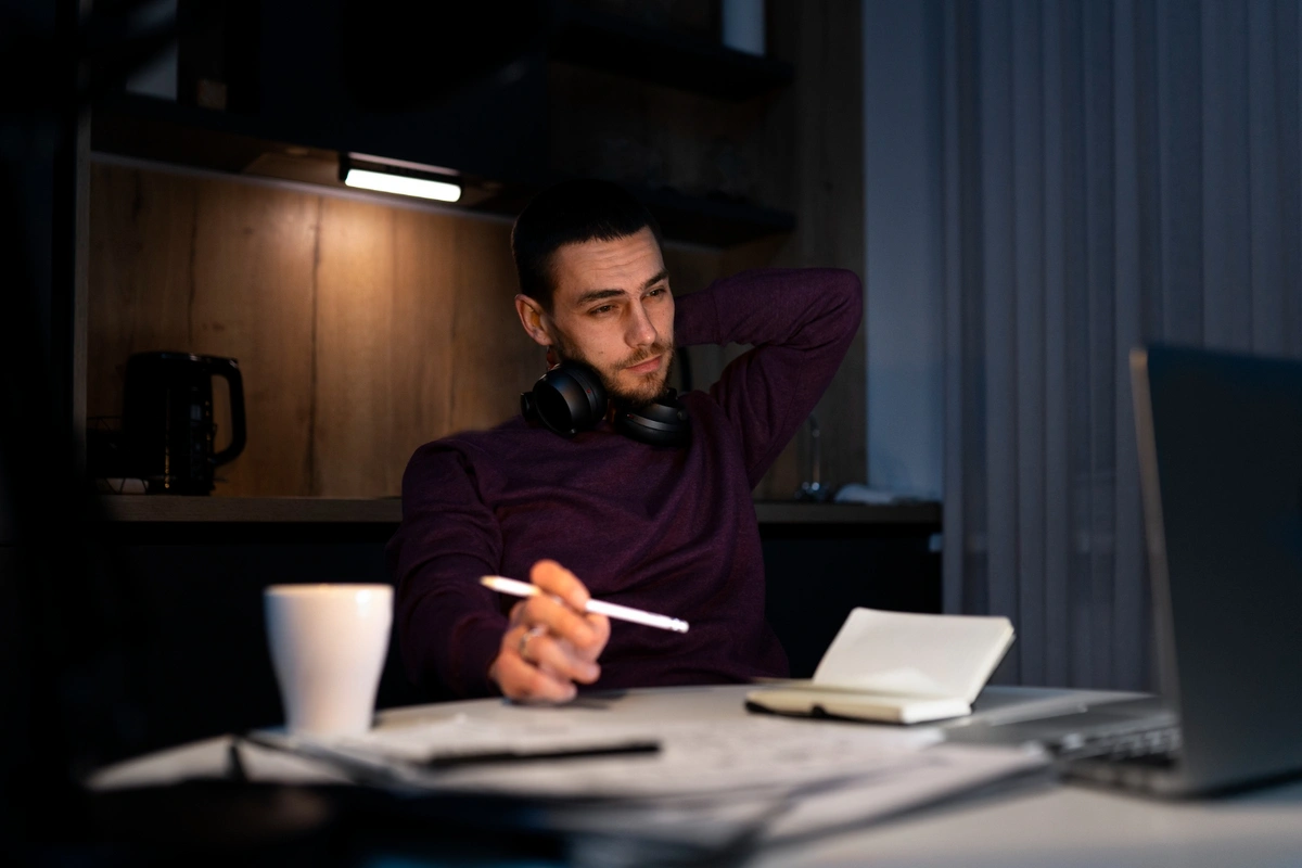 A man in a casual sweater sits at a desk in a softly lit room, looking thoughtful with headphones around his neck. He holds a pen in one hand, with an open notebook and a laptop on the table, suggesting he is engaged in late-night work or planning.