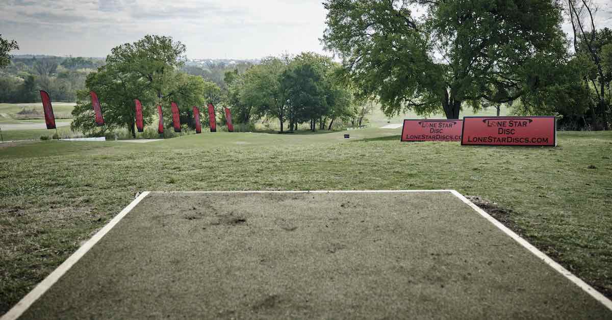 A turf disc golf tee pad in an open, mown area with scattered mature trees