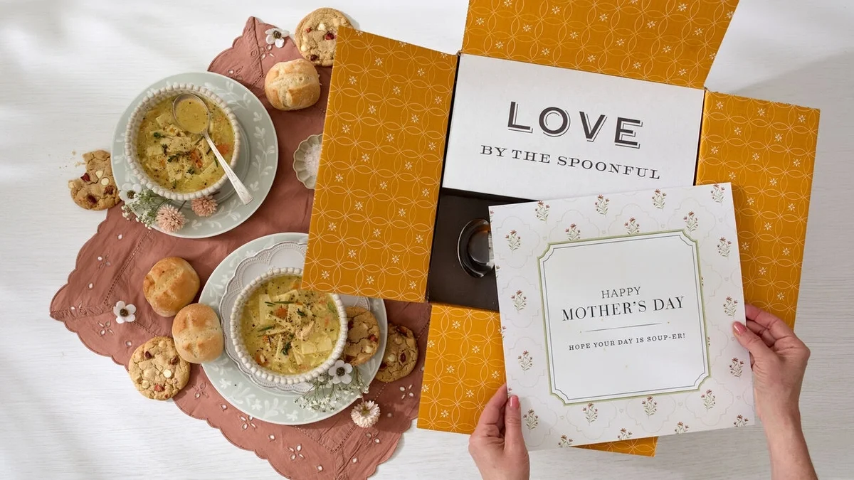 Overhead view of a Mother’s Day gift arrangement with two bowls of chicken soup, rolls, cookies, and an open yellow Spoonful of Comfort gift box. Hands hold a card that reads “Happy Mother’s Day,” with a “Love by the Spoonful” card inside the box.