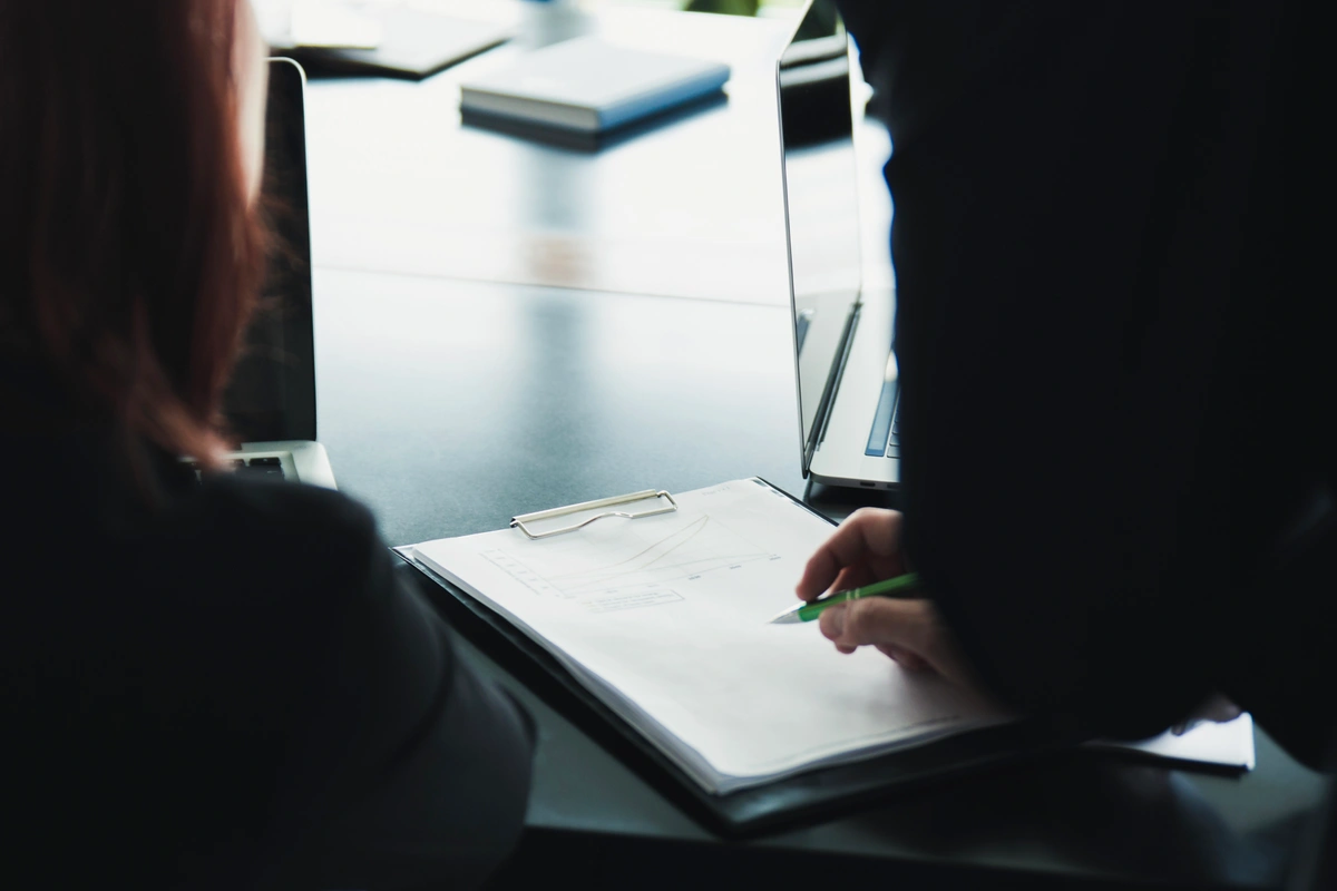 Businesspeople reviewing documents on a clipboard during a meeting, with laptops on the table.