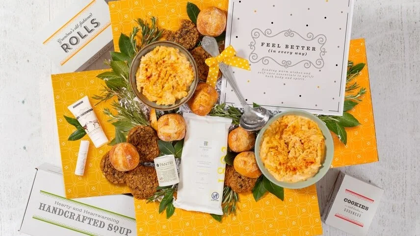 Overhead view of a care package arrangement featuring bowls of macaroni and cheese, rolls, cookies, soup packaging, wellness items, greenery, and a “Feel Better” card displayed on a yellow patterned background.