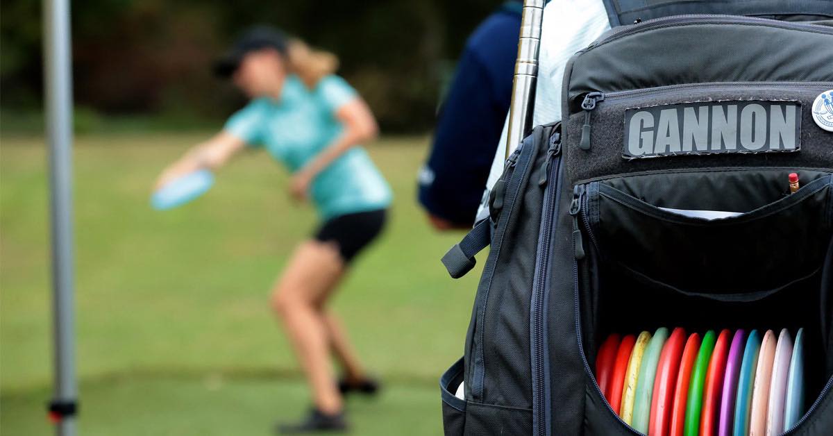 Close-up of a disc golf bag with the last name "Gannon" on it. Blurred image of Missy Gannon throwing in background