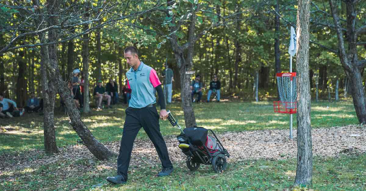 A young man with a disc golf cart walks away from a red disc golf basket among a small group of trees in an otherwise open field