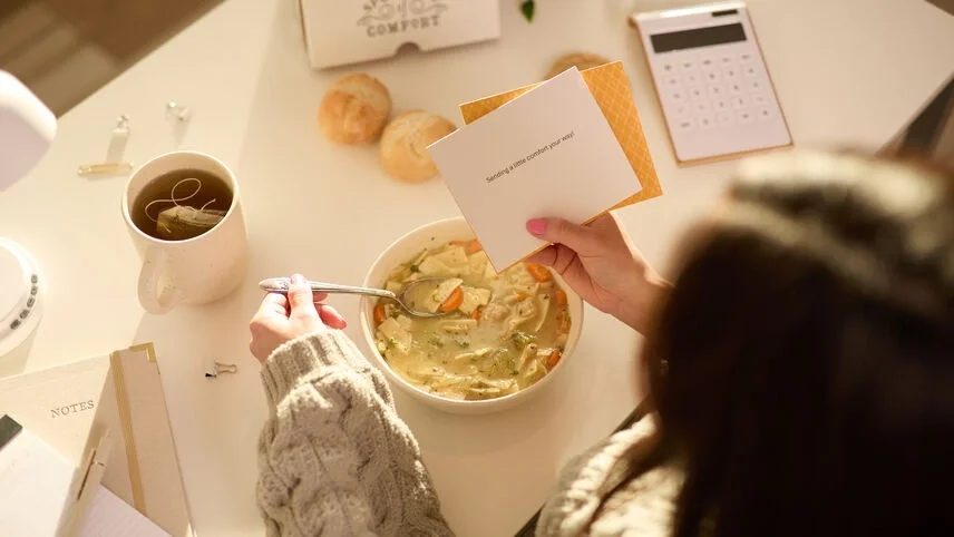 Overhead view of a person in a knit sweater eating a bowl of chicken soup while holding a handwritten note, with a mug of tea, rolls, and a calculator on the table nearby.