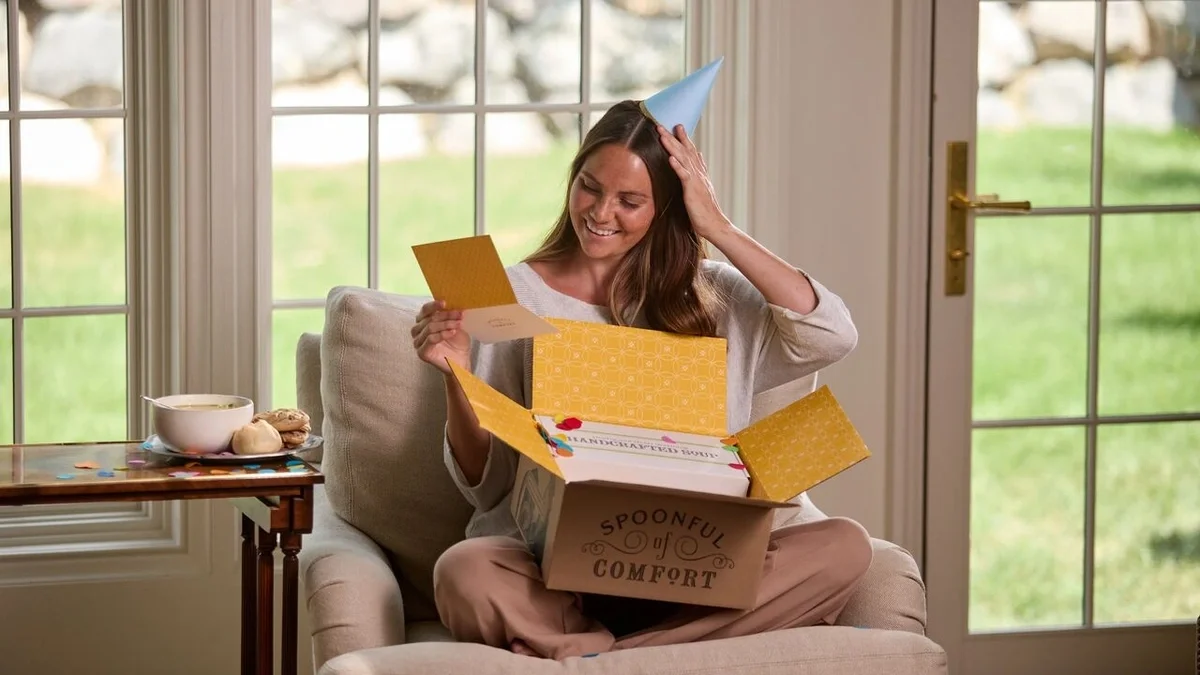 Woman smiles as she places a birthday party hat on her head, and reads a card from an opened Spoonful of Comfort birthday package